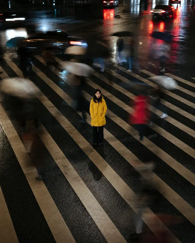 城市斑马线动态人像:一位穿浅色风衣的人站在城市斑马线中央,身体稳定,周围行人形成动态模糊,繁忙路口,信号灯和车灯形成背景色块,不出现可读广告牌。